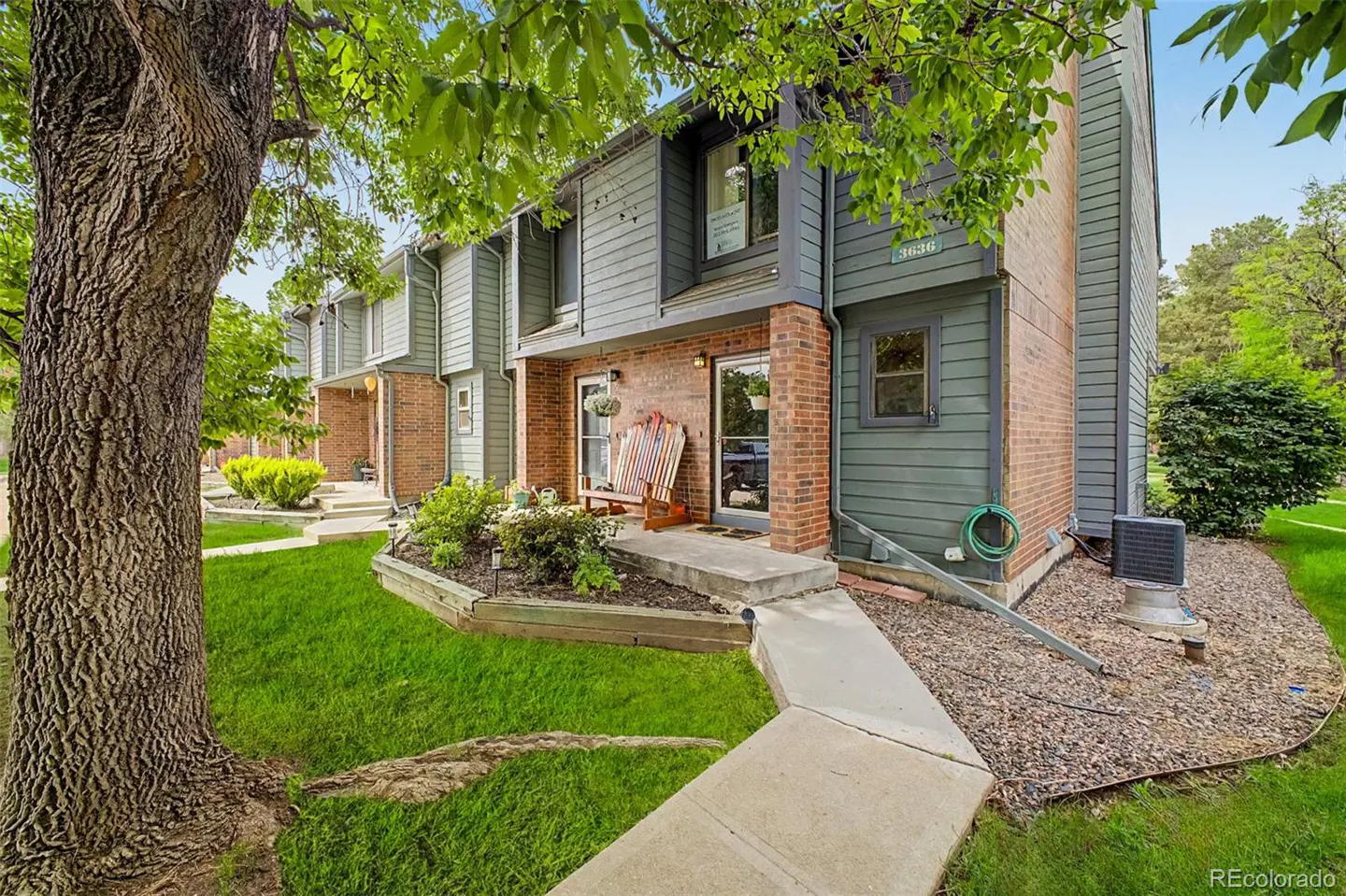 Exterior view of a two-story townhouse with brick and green siding, a concrete walkway, and a tree in the front yard.