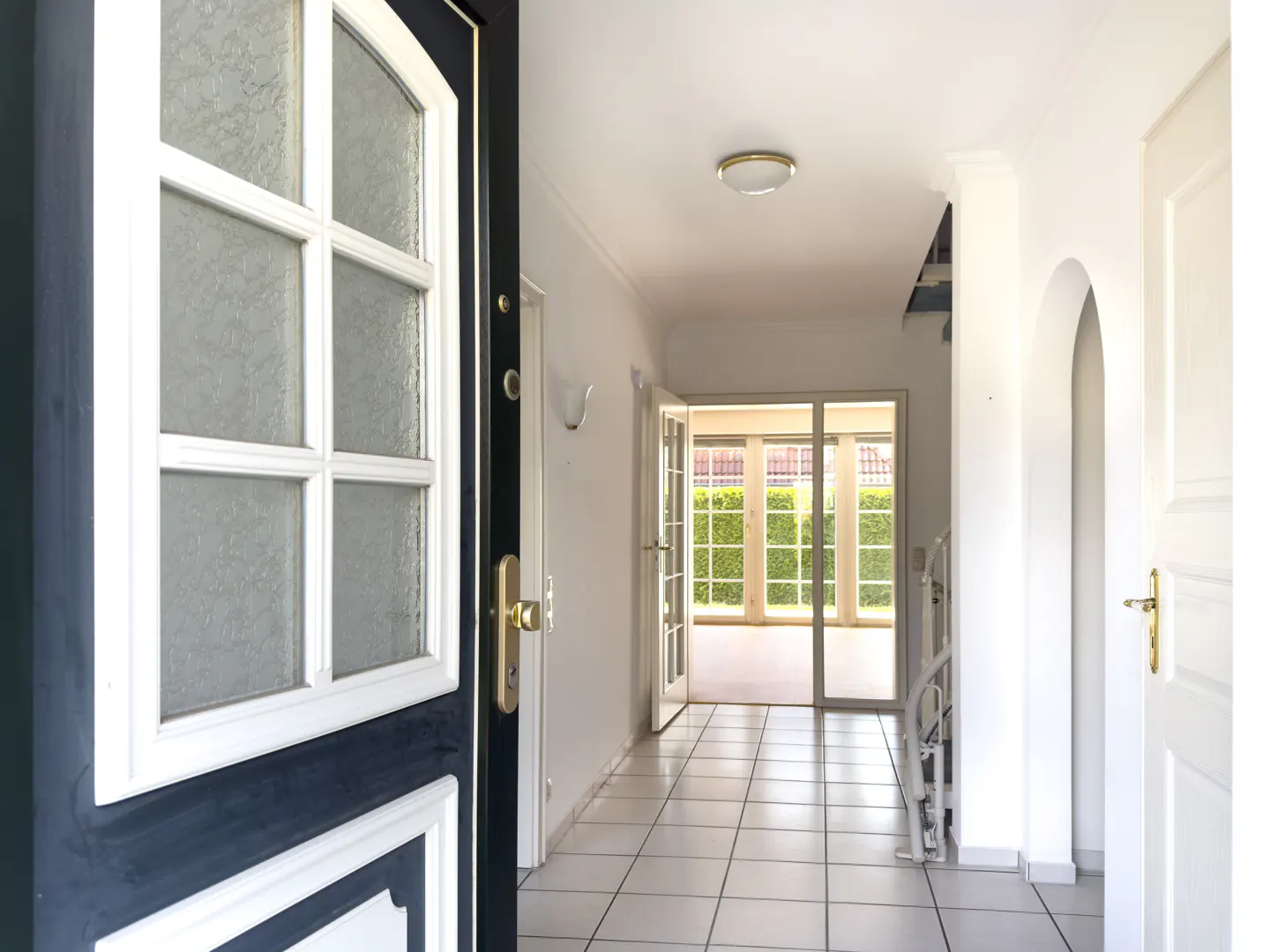 Open front door with glass panes leads into a bright hallway with white tile floor and a view of a sunroom.