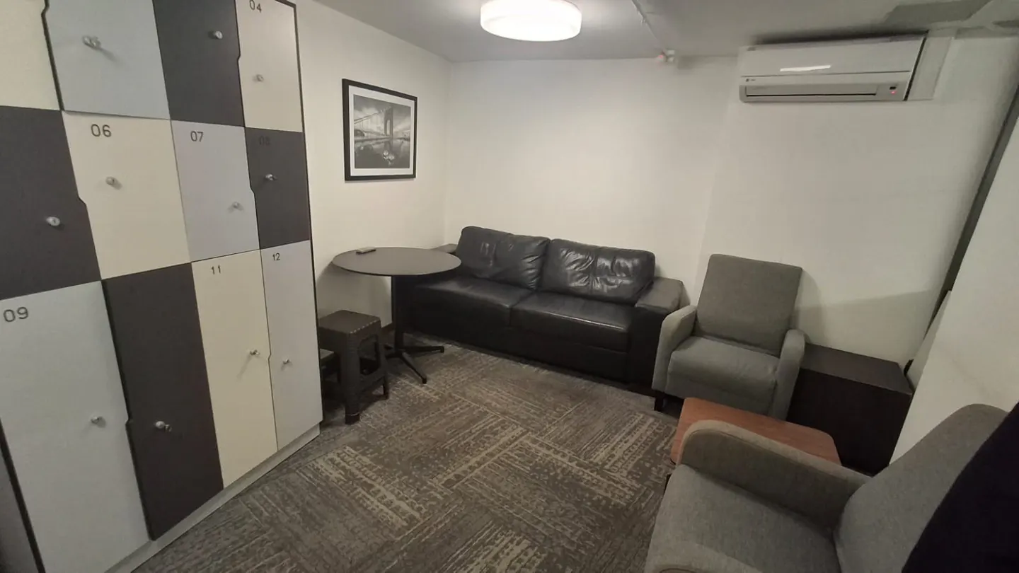 A small waiting room with lockers, a black leather couch, and two gray armchairs. A small table and stool are near the lockers.
