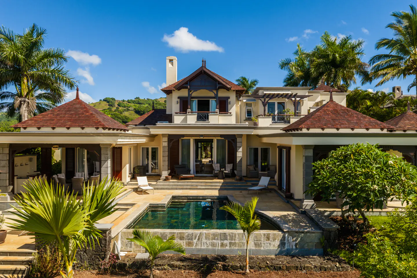 Luxury villa with a pool, surrounded by palm trees under a blue sky. The villa has a brown roof and a beige exterior.