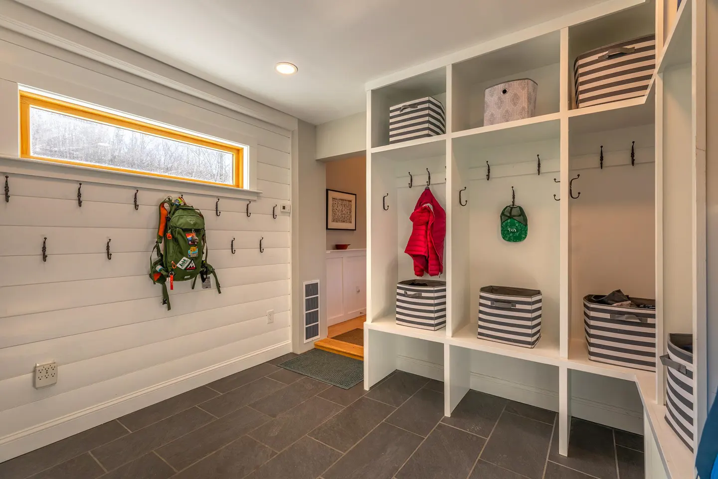 Mudroom with white shiplap walls, dark tile floor, and built-in white cubbies with hooks and striped storage boxes. A window is above hooks with a backpack.