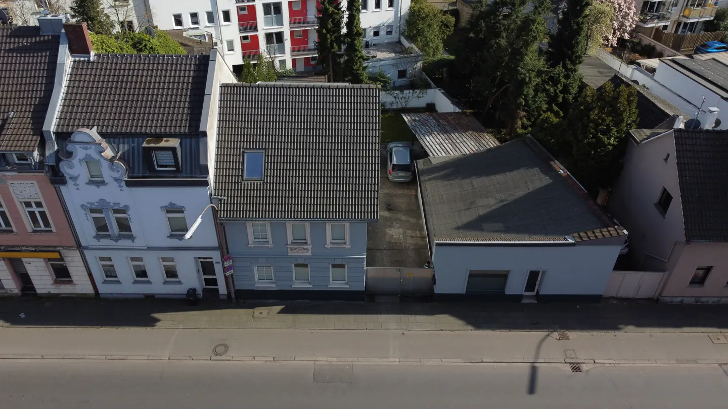 Aerial view of a light blue two-story house with a gray tiled roof, next to a white building and a gray garage. A car is parked in the driveway.
