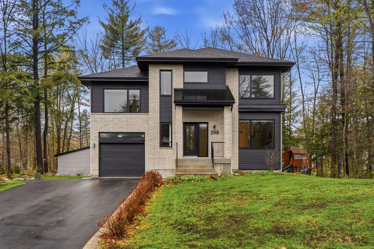 Two-story modern home with light brick and dark gray siding, black trim, and a black garage door, surrounded by trees and a green lawn.