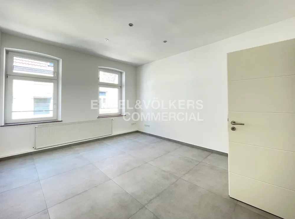 Bright, empty room with gray tile floor, white walls, and two windows. A white door is slightly ajar on the right. Radiators are under the windows.