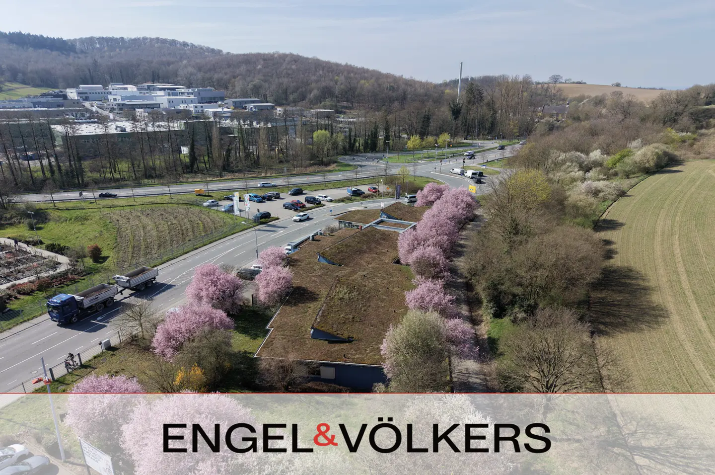 Aerial view of a building with a green roof, pink flowering trees, and a road with cars and trucks.