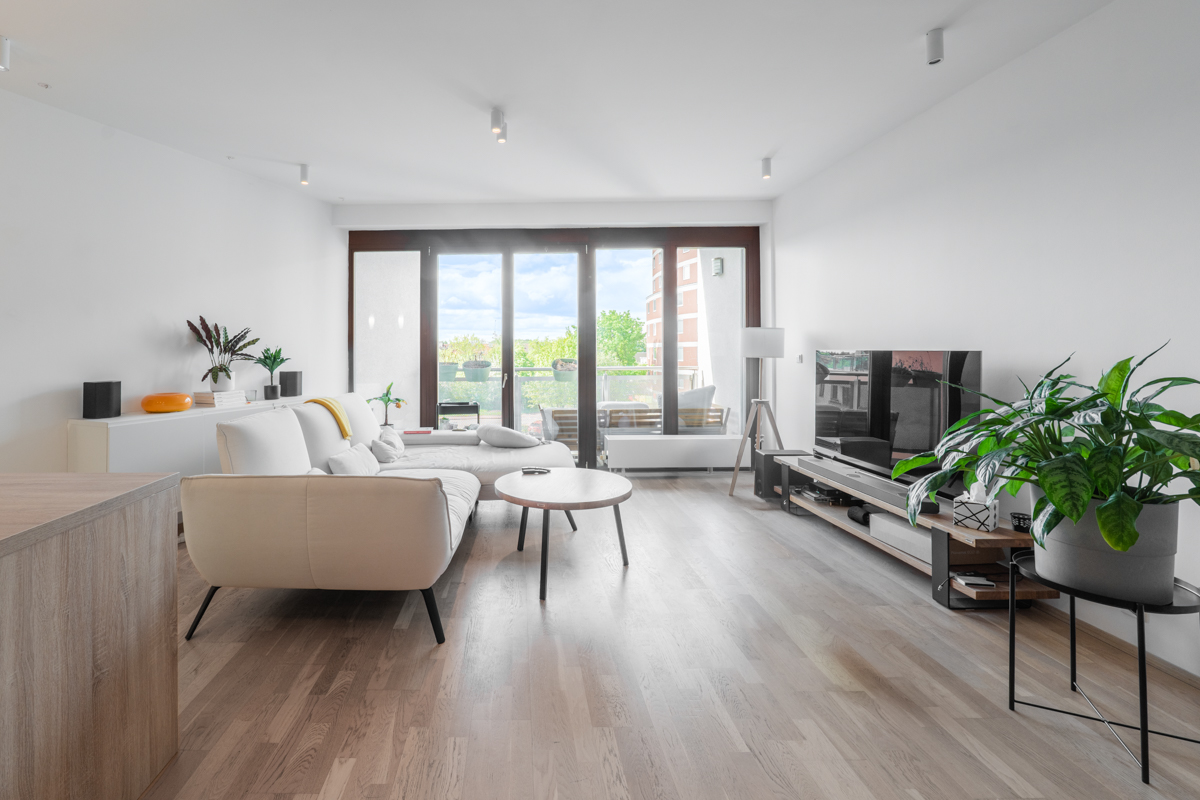 Bright living room with white walls, wood floors, and a large sliding glass door to a balcony. A white sofa and TV stand are visible.