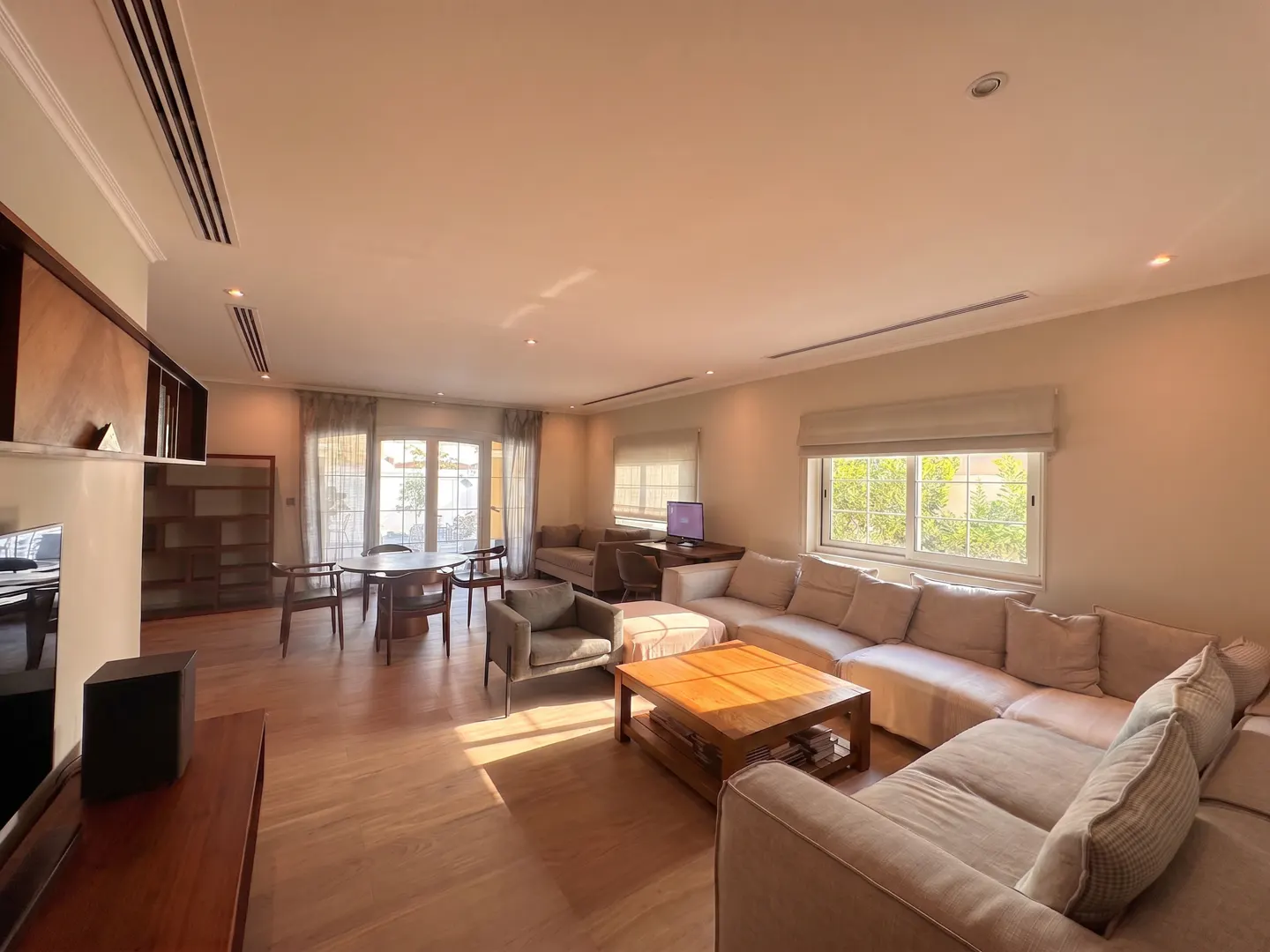 A bright, open living room with a large beige sectional sofa, wooden coffee table, dining table, and natural light.