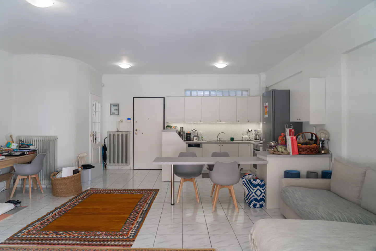 Open-plan kitchen and living area with white cabinets, a grey dining set, tiled floors, and a patterned area rug.