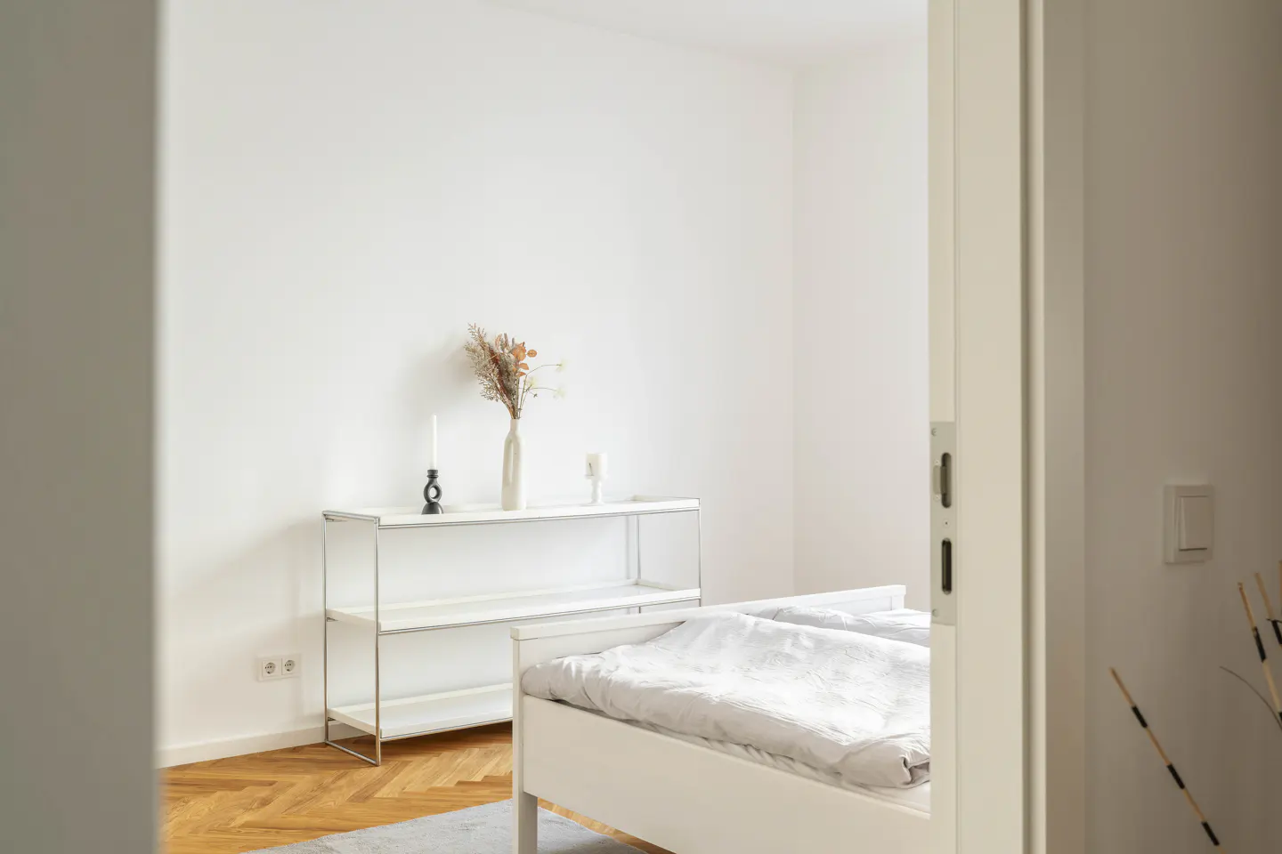 A bright bedroom with white walls, a white bed, and a white shelf with a vase of dried flowers.