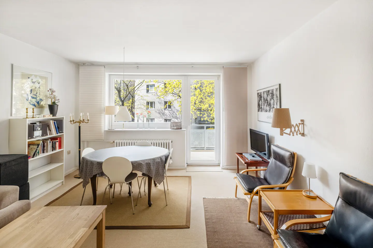 Bright living room with white walls, a round table with chairs, and a large window overlooking a building and trees.