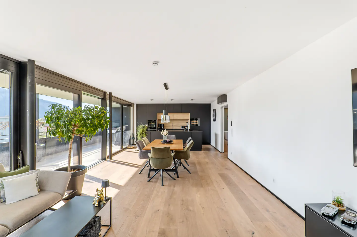 Bright, modern apartment interior with wood floors, a dining table with six chairs, and a black kitchen. Large windows offer natural light.
