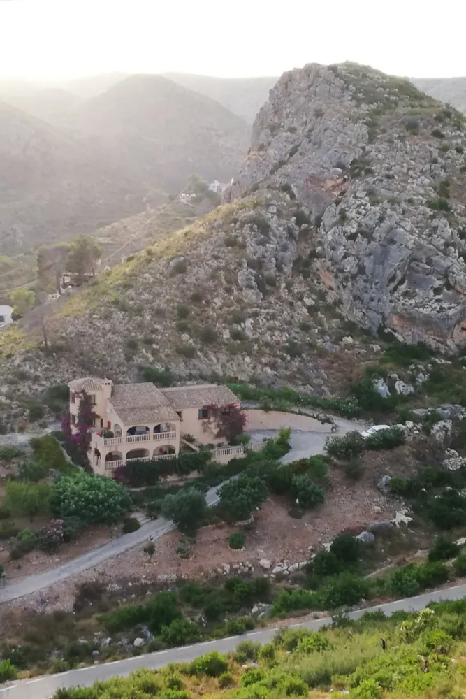 A tan house with a tile roof sits on a hillside with a large rock formation behind it.
