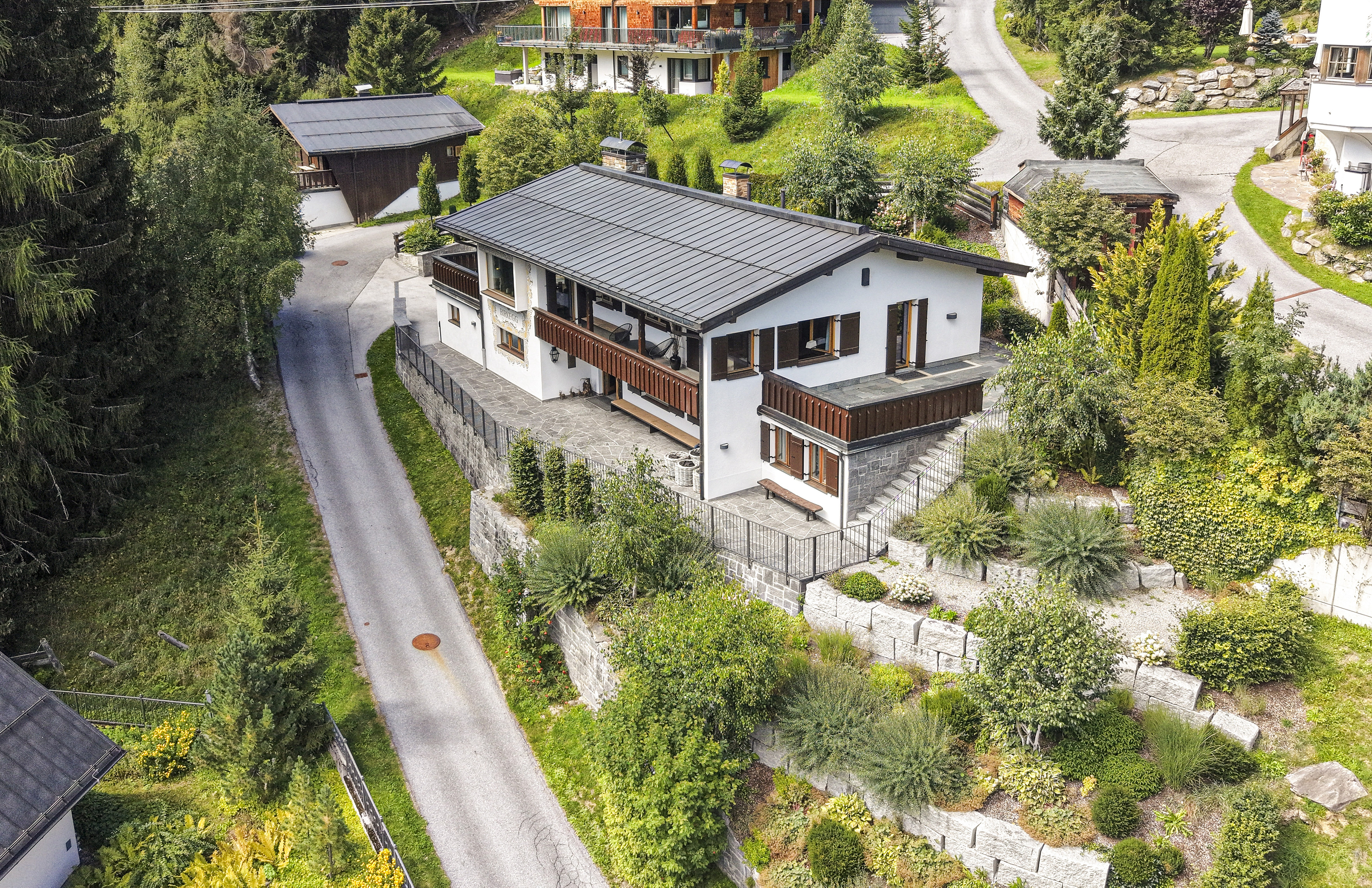 Aerial view of a white chalet-style house with a dark roof, wooden balconies, and stone landscaping in a lush, hilly area. [AI-generated text]