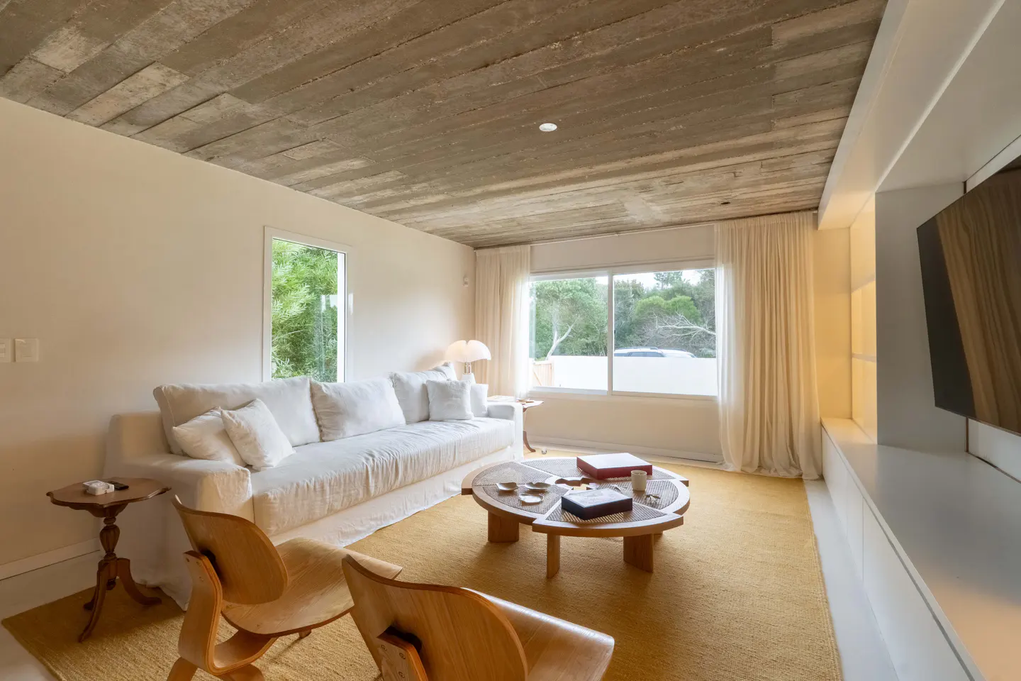 Living room with a concrete ceiling, white walls, and a yellow rug. A white sofa and wooden chairs are arranged around a round coffee table. Natural light streams through the windows.