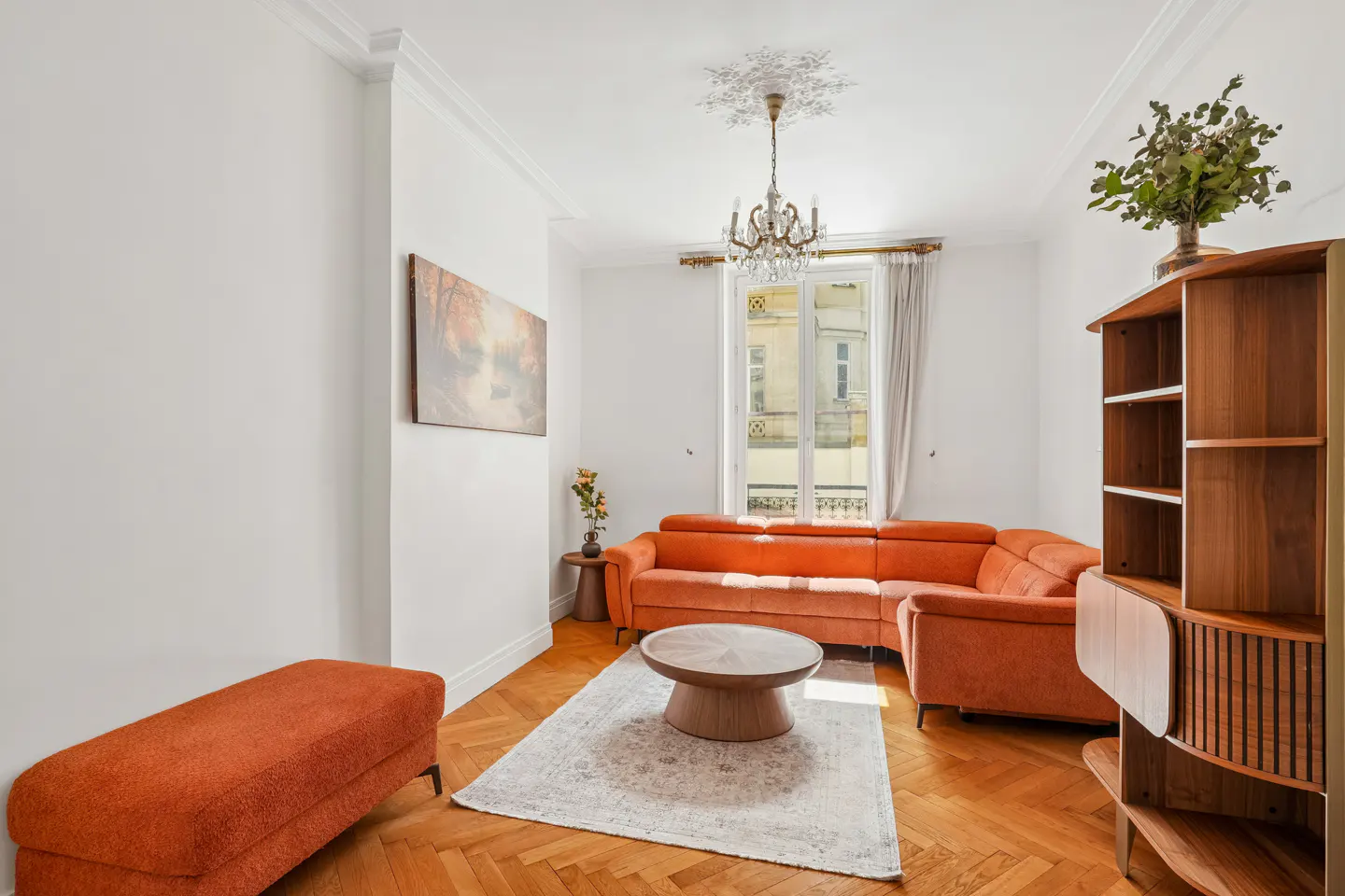 Bright living room with orange sectional sofa, round wood table on a patterned rug, and a wood bookshelf. A chandelier hangs from the ceiling.