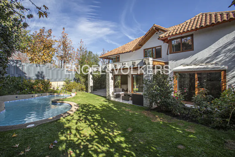 Backyard of a two-story home with a swimming pool, green lawn, stone patio, and terracotta roof under a blue sky.