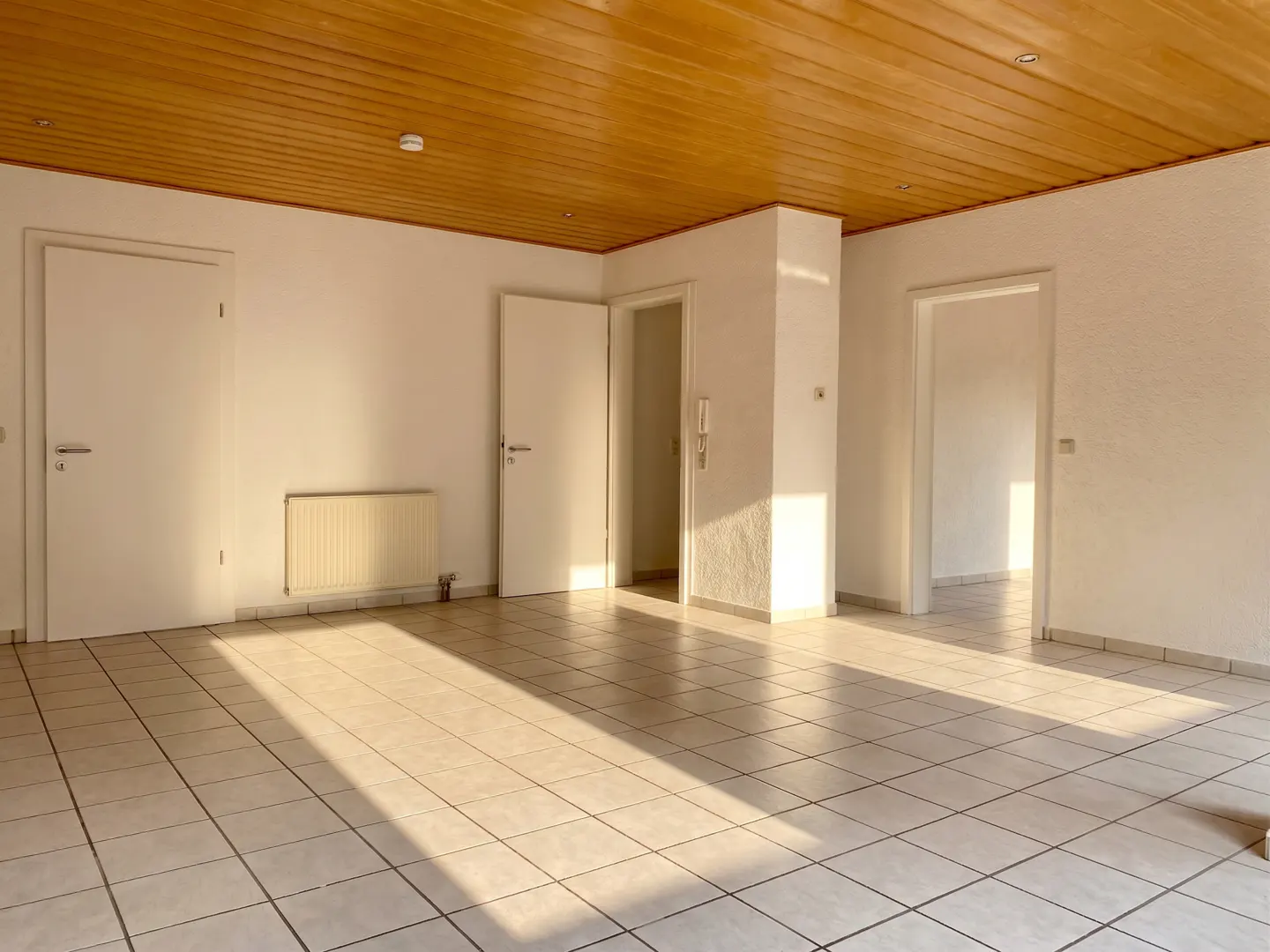 Empty room with white tiled floors, white walls, multiple doorways, and a warm-toned wooden paneled ceiling.
