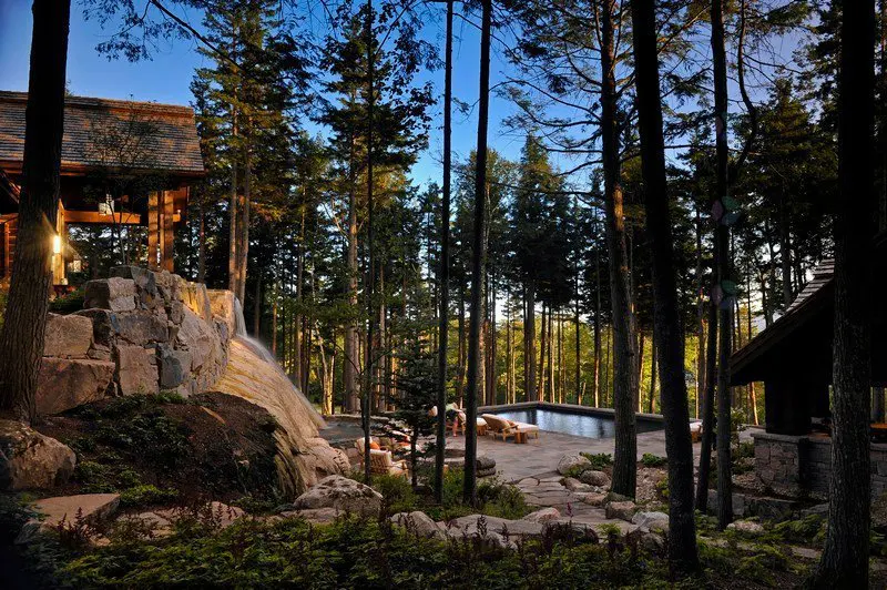 Estate backyard with pool. Tall trees surround a stone patio with lounge chairs. A waterfall cascades down a rock formation.