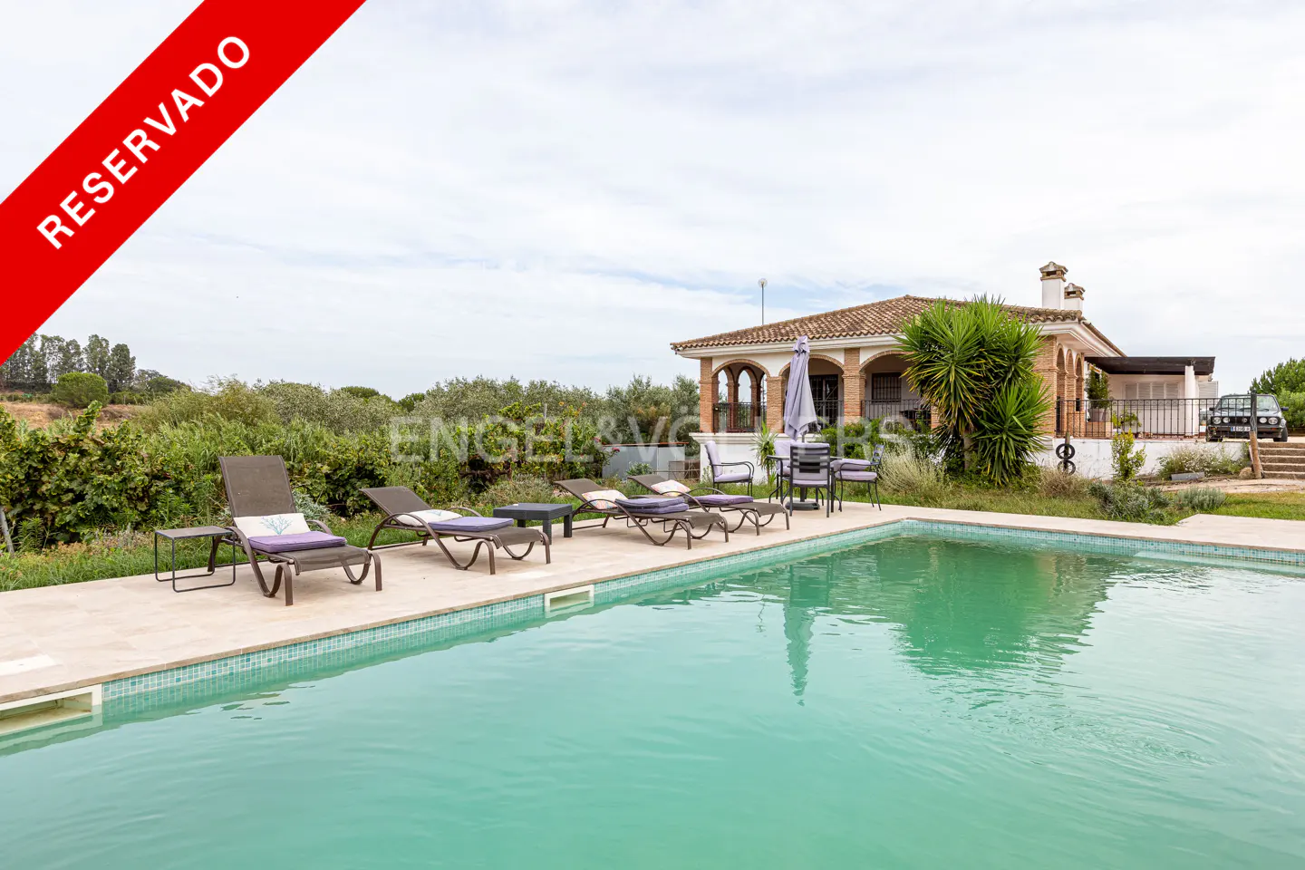 Reserved property with a pool. Lounge chairs sit poolside, with a house featuring arched windows and a red tile roof in the background.