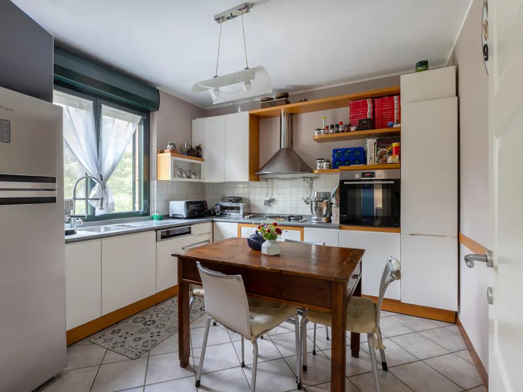 Bright kitchen with white cabinets, stainless steel appliances, and a wooden table with chairs. A window with sheer curtains lets in natural light.