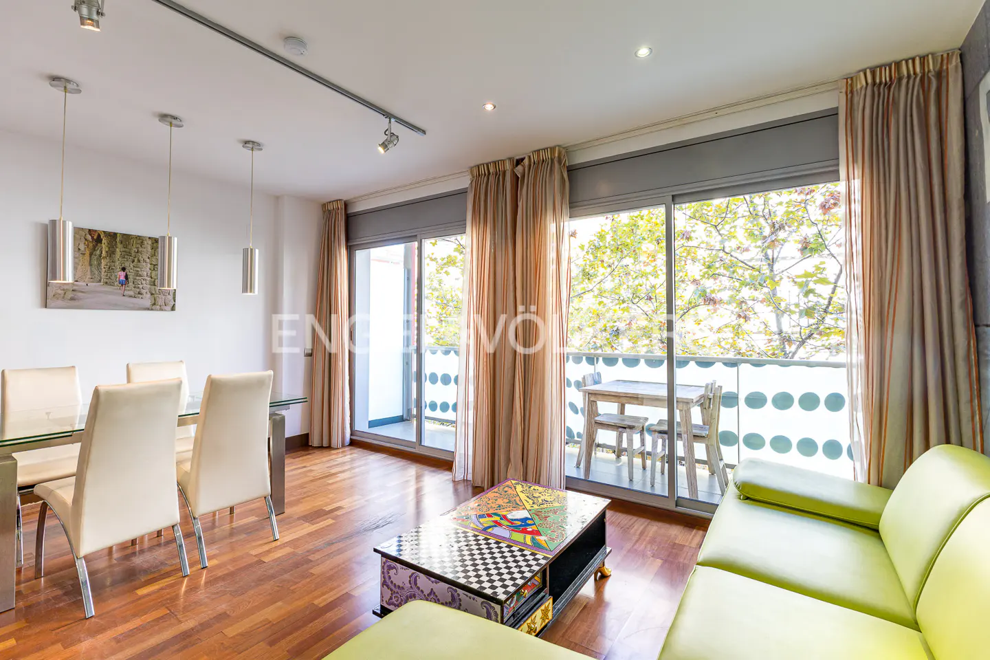 Bright living room with wood floors, a green sofa, and a glass dining table with cream-colored chairs. Balcony with table and chairs visible through sliding glass doors.