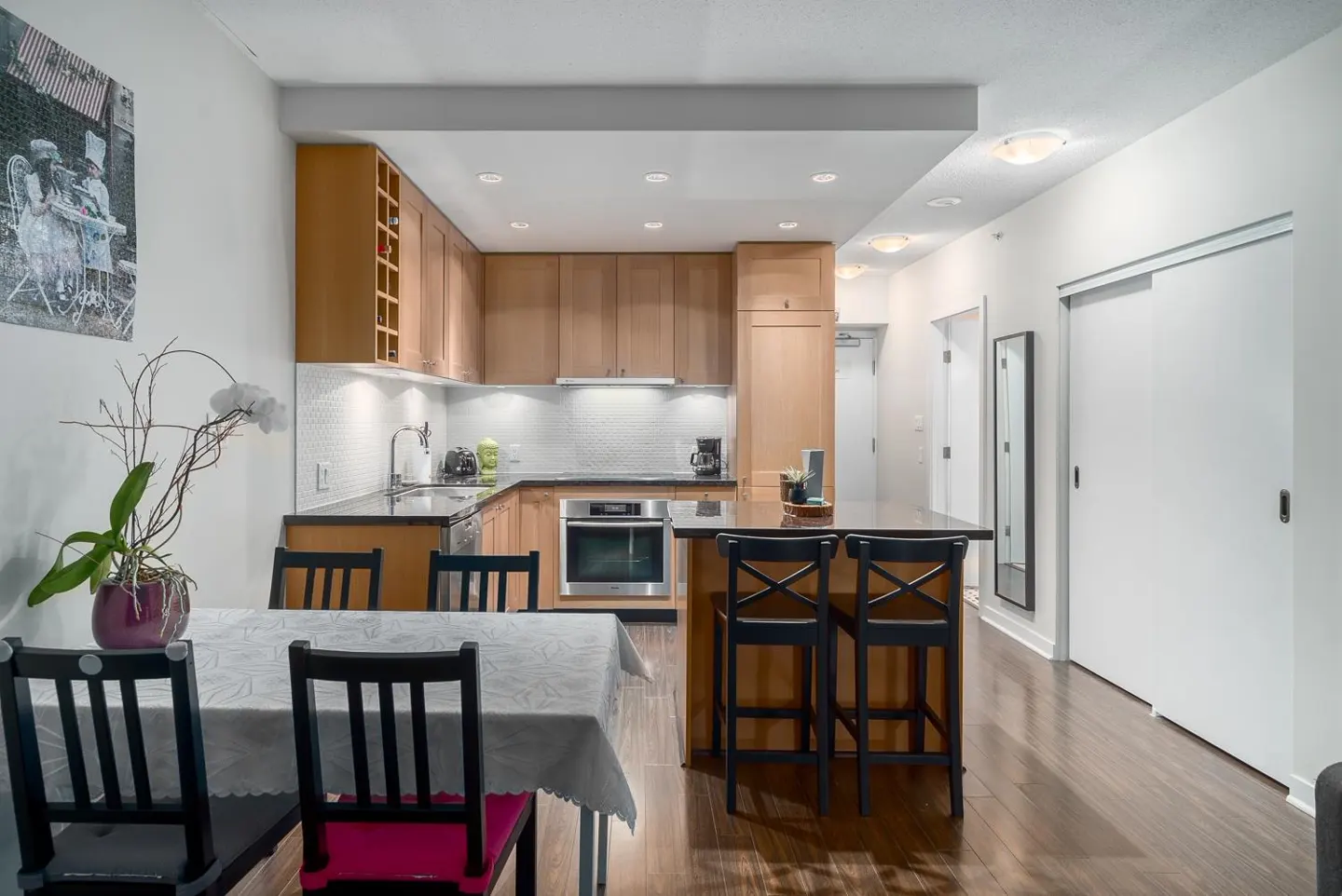 A bright kitchen with wood cabinets, black countertops, and a dining table with black chairs. A kitchen island with bar stools is also visible.