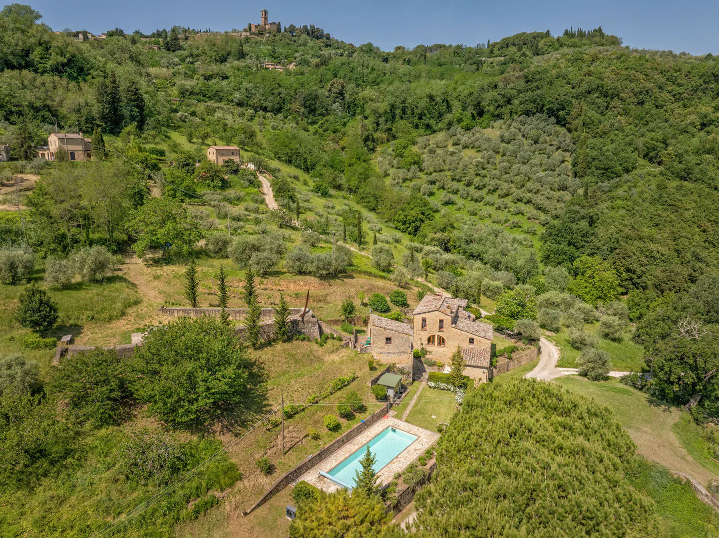 Aerial view of a stone villa with a swimming pool, nestled in a lush, hilly Tuscan landscape with olive groves.