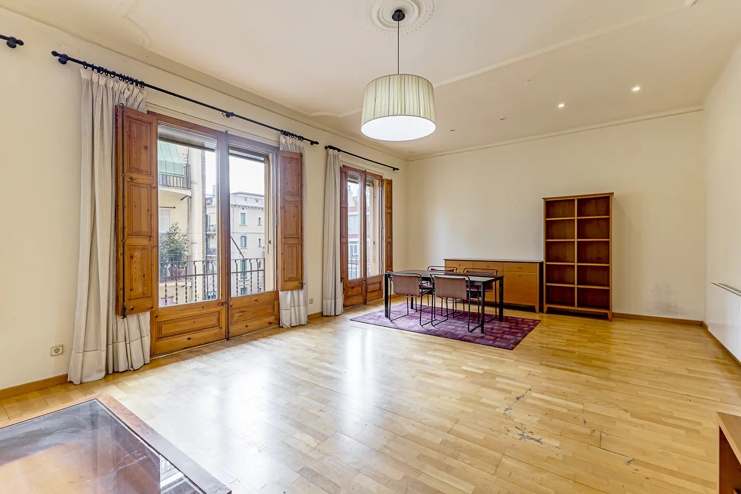 Bright, airy room with wood floors, shutters, and balcony views. Dining table with four chairs sits on a patterned rug. A bookshelf and cabinet are against the wall.