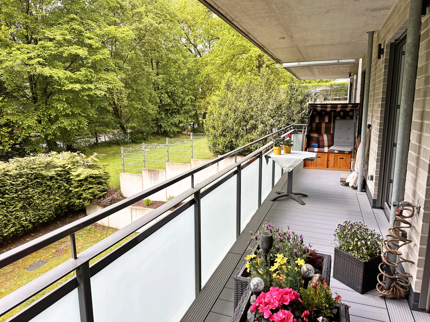 Balcony with gray flooring, glass railings, and potted flowers. A table, beach chair, and green trees are in the background.