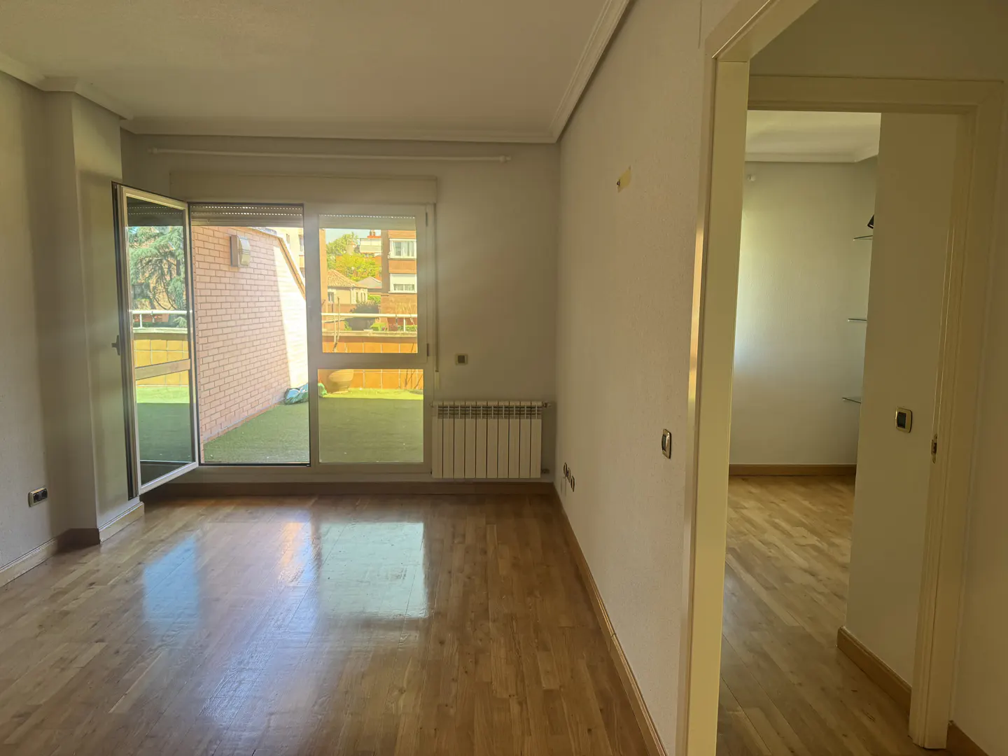 Bright, empty living room with hardwood floors, white walls, and a sliding glass door to a balcony. A doorway leads to another room.