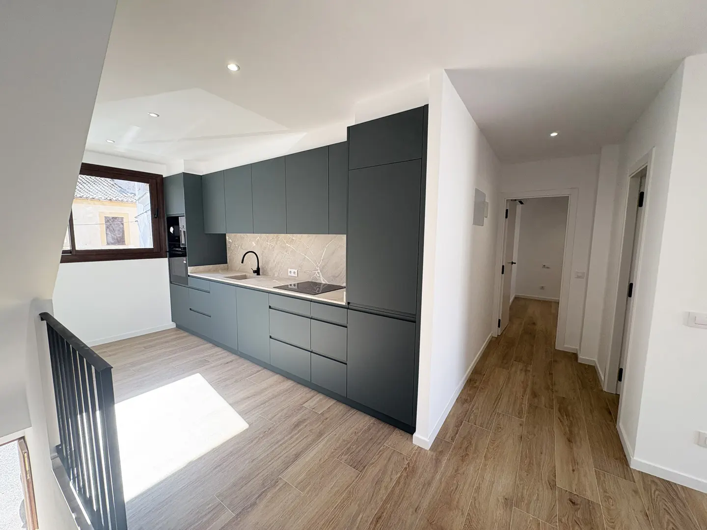 A modern kitchen with gray cabinets, marble backsplash, and wood-look tile flooring. A hallway with white walls and doors is visible.