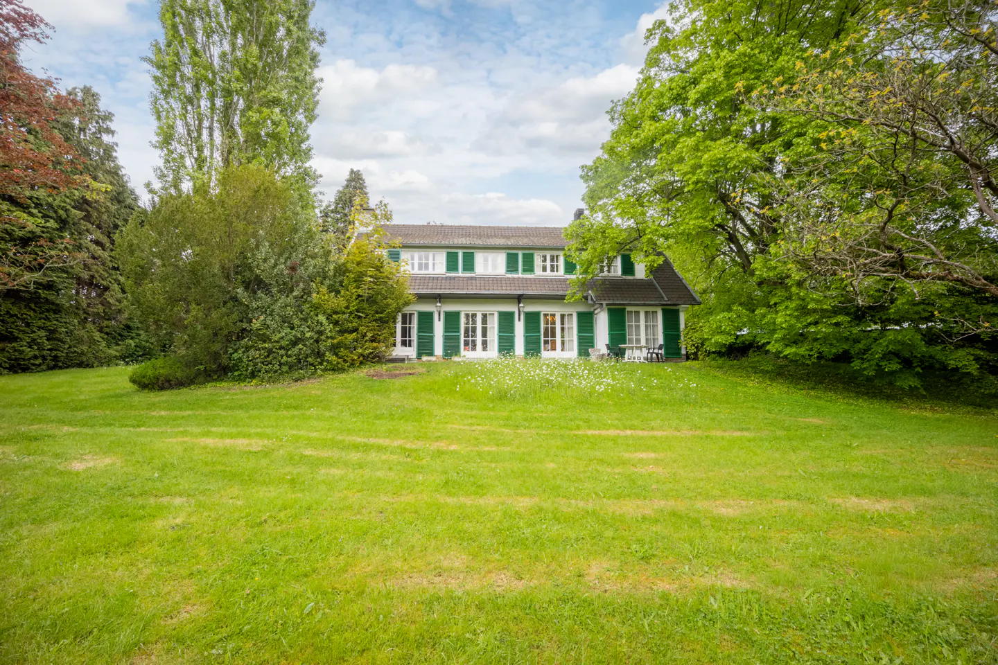 Two-story house with green shutters and a large lawn, framed by trees under a cloudy sky.