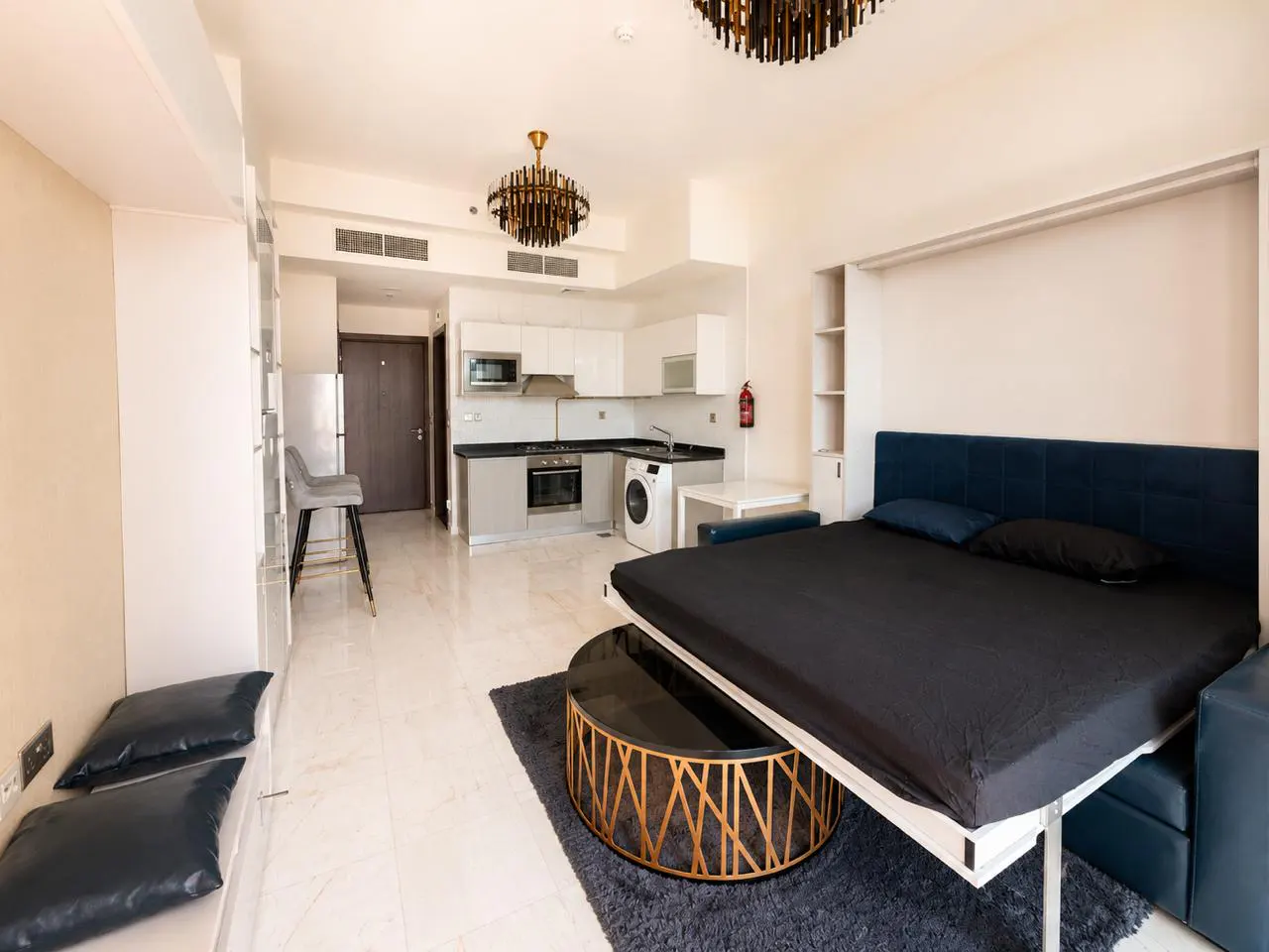 Studio apartment with a blue Murphy bed, black bedding, and a gold coffee table. The kitchen has white cabinets and gray countertops.