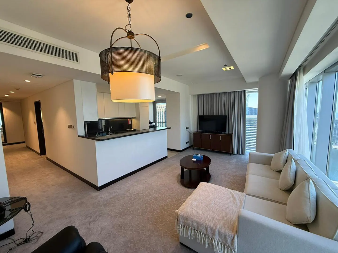 A modern apartment interior with a white sofa, round table, TV, and kitchen counter. A decorative chandelier hangs above.