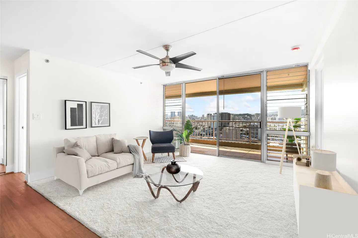 Bright living room with white sofa, glass coffee table, dark armchair, and floor-to-ceiling windows overlooking a city.