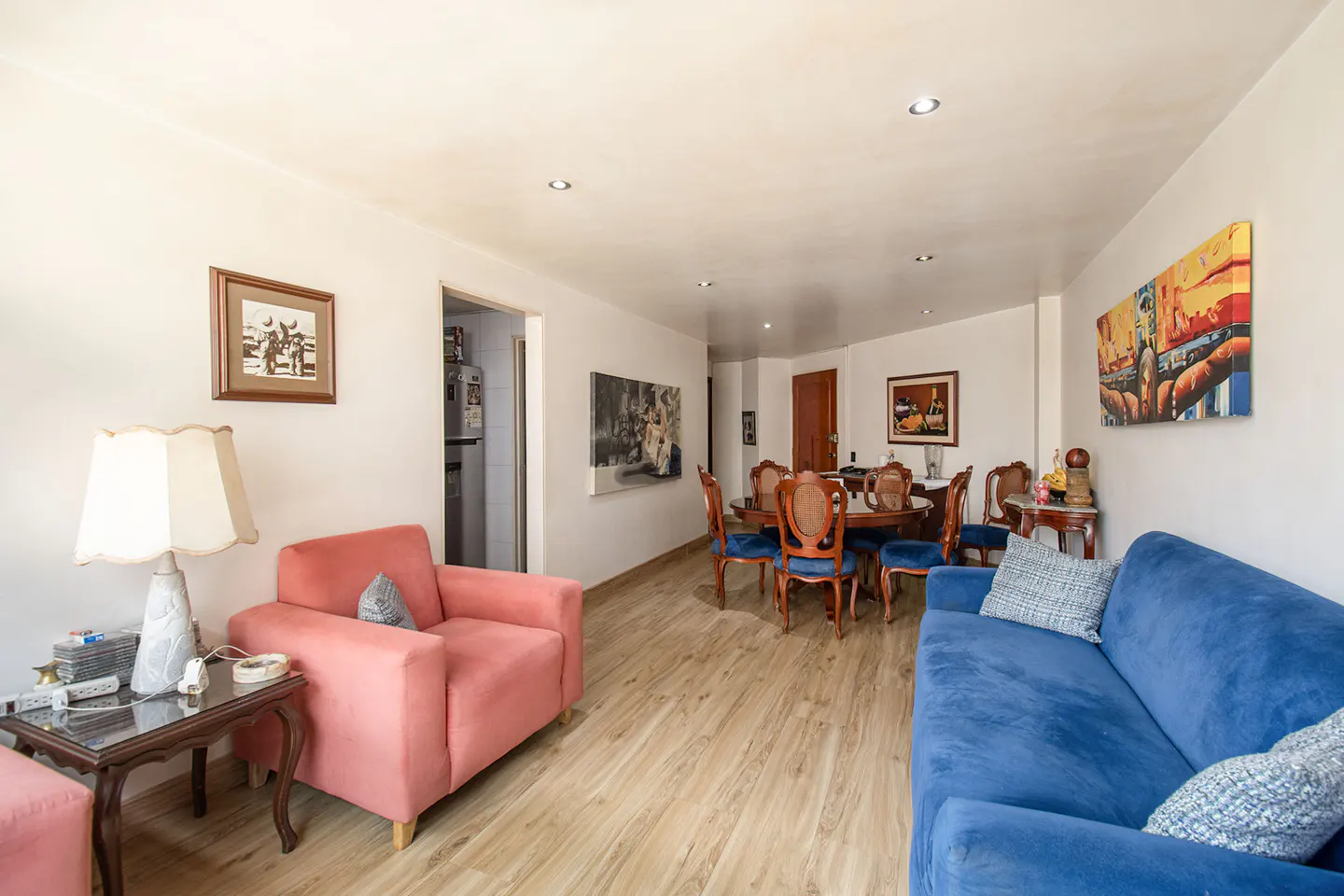 Bright living room with wood floors, a blue sofa, a pink armchair, and a dining table with blue chairs. Artwork adorns the white walls.