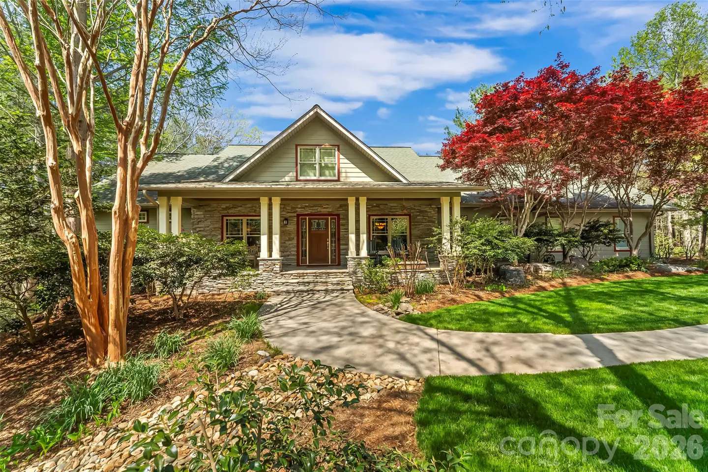 A single-story stone house with a green roof, white columns, and a red maple tree on a sunny day.