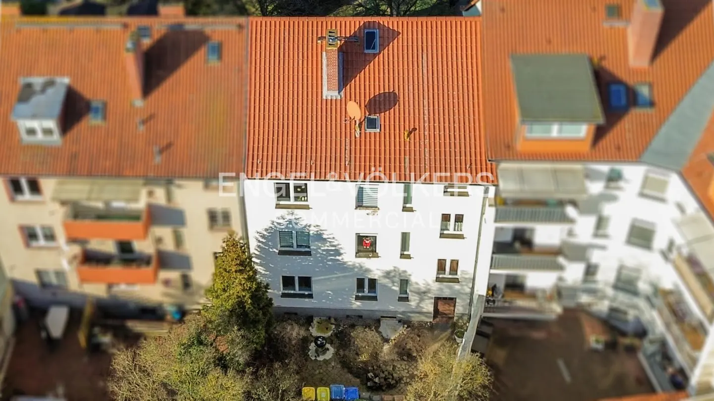 Aerial view of a white building with a red tile roof, chimney, and satellite dish, surrounded by other buildings and trees.