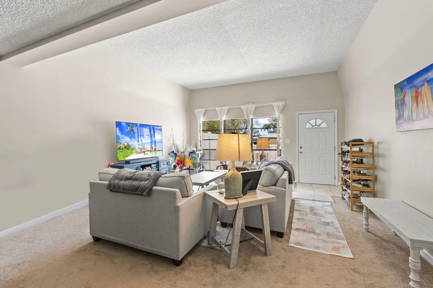 Living room with beige walls, carpet, and furniture. A TV displays a beach scene. A white front door and window are visible.