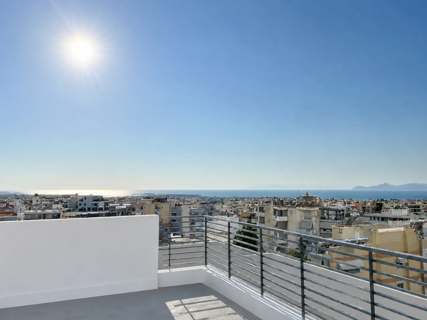 View from a rooftop terrace with metal railings overlooking a city with the ocean in the distance under a bright blue sky.