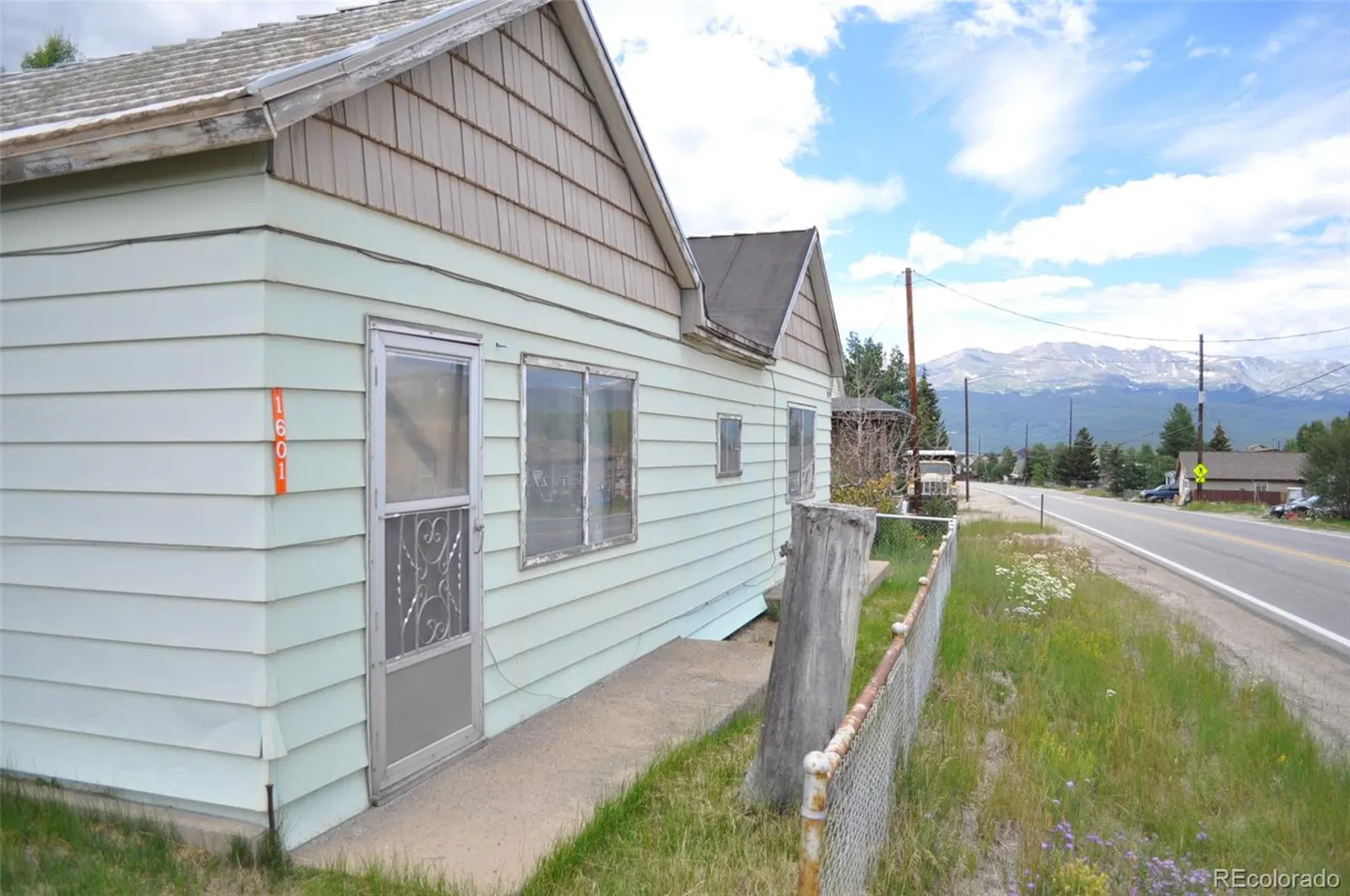 Exterior view of a light green house with a screen door, windows, and a brown shingled roof. Mountains are visible in the background.