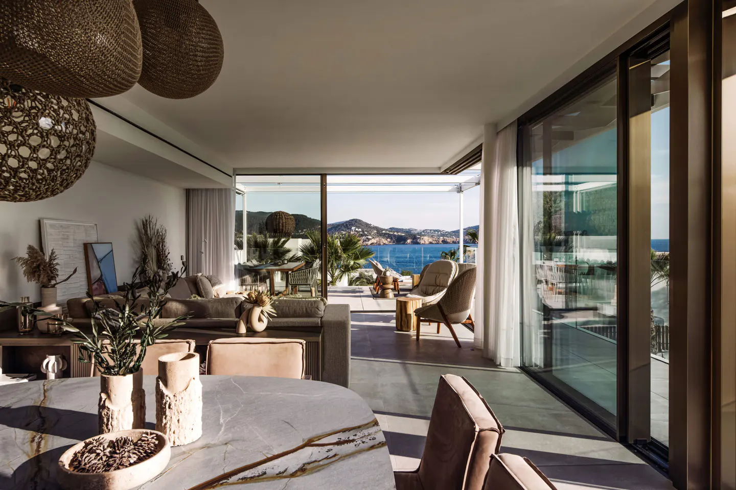 Bright, modern living room with a view of the ocean. Wicker light fixtures hang above a stone table and chairs. Sliding glass doors open to a patio.