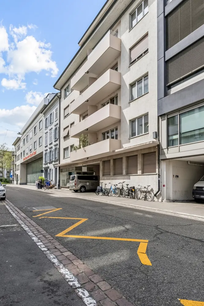 Street view of a modern, multi-story white apartment building with balconies, parking garage, and bicycles parked outside. Yellow road markings are visible.