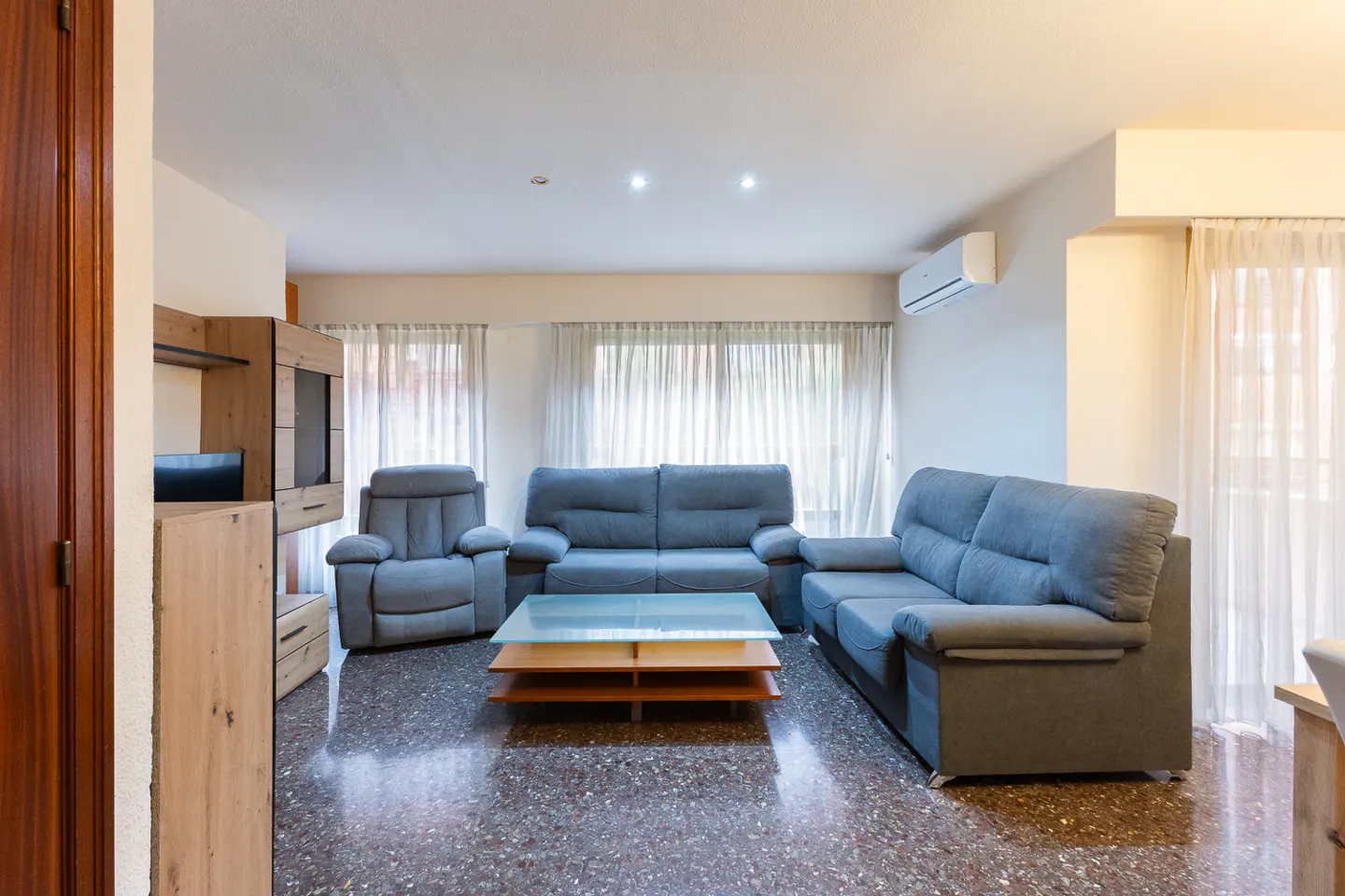 Living room with blue sofas, glass coffee table, and wood cabinet. Windows with sheer curtains let in natural light.