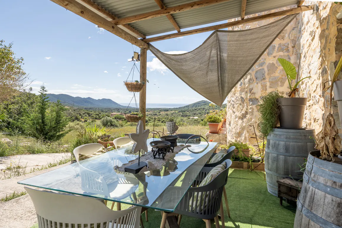 Outdoor dining area with a glass table, chairs, and decorative items under a wooden pergola with a view of green hills.