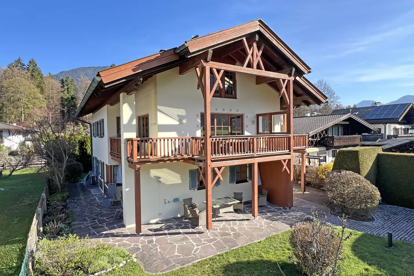 Two-story house with brown trim and balconies, surrounded by green grass and trees. A stone patio with outdoor furniture is visible.