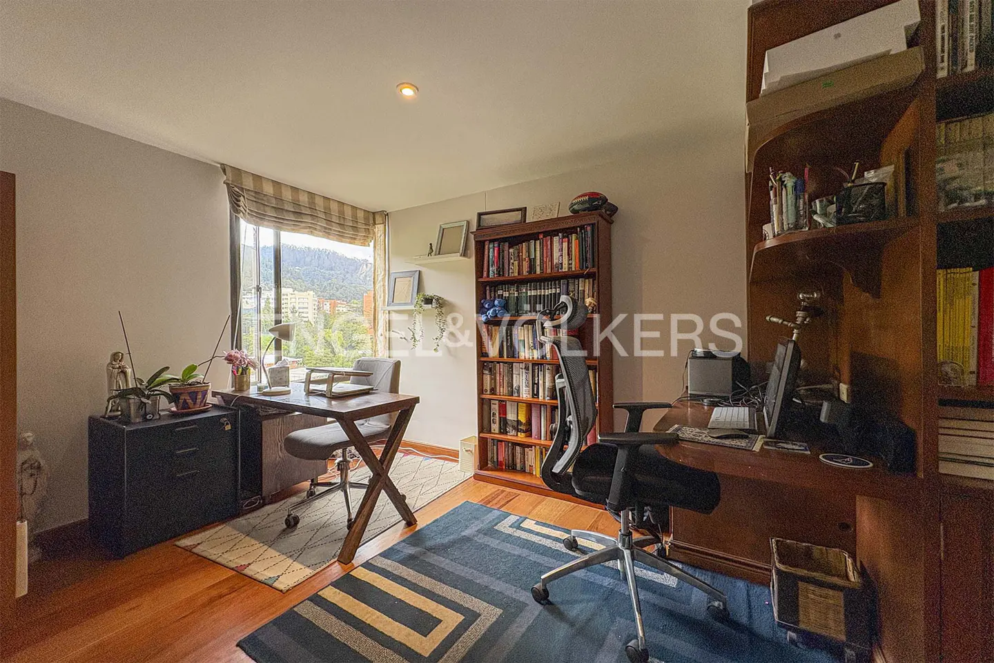 A home office with a wooden floor, a desk, a chair, a bookcase, and a window with a view of a mountain.