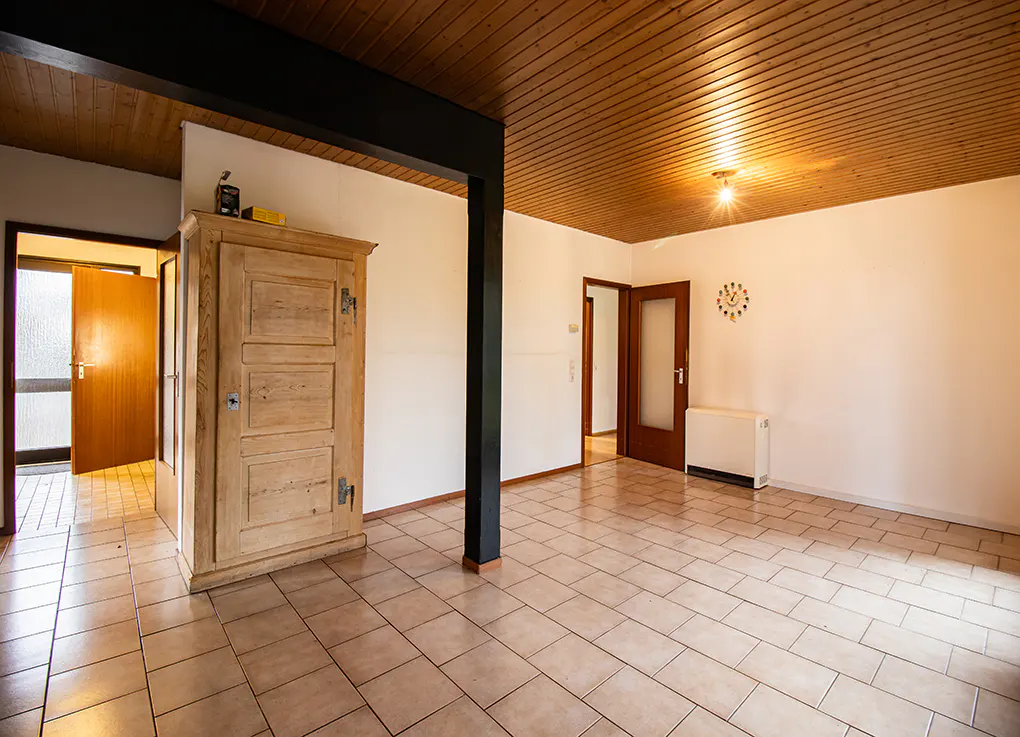A bright room with beige tile flooring, a wood ceiling, and white walls. A wooden cabinet stands near an open doorway.