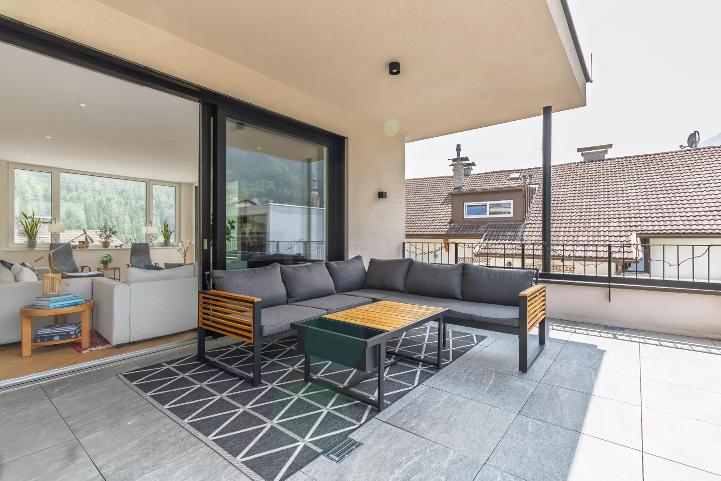 A modern balcony with gray sectional sofa, wood table, and patterned rug. Sliding glass doors open to a living room with mountain views.