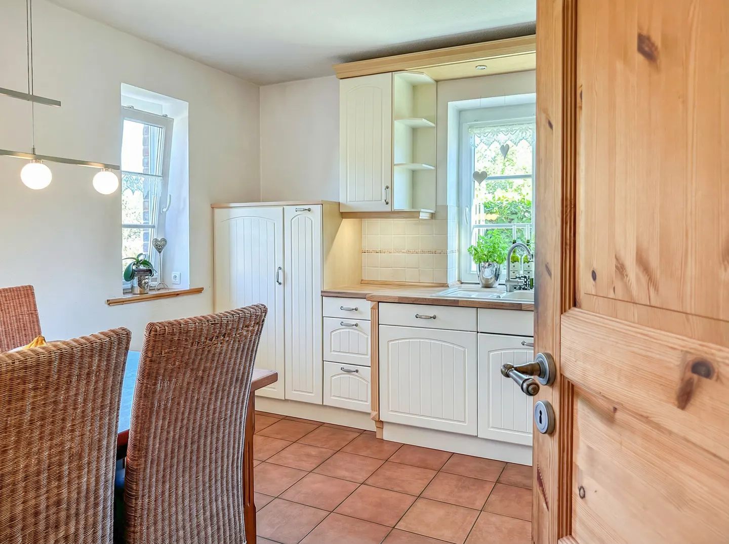 A bright kitchen with white cabinets, a wood countertop, and a window with a heart-shaped curtain. A wooden door is ajar.